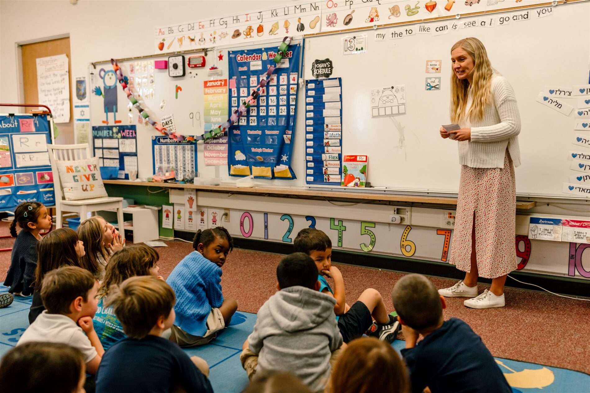 A teacher at Kavod Charter School stands in front of a class of young children. The children are sitting on a colorful carpet and wearing uniforms. The classroom is brightly decorated.