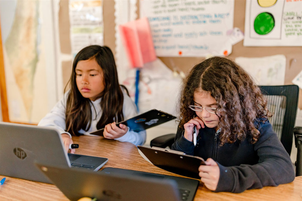 Two Kavod students hold clipboards and work on laptops.