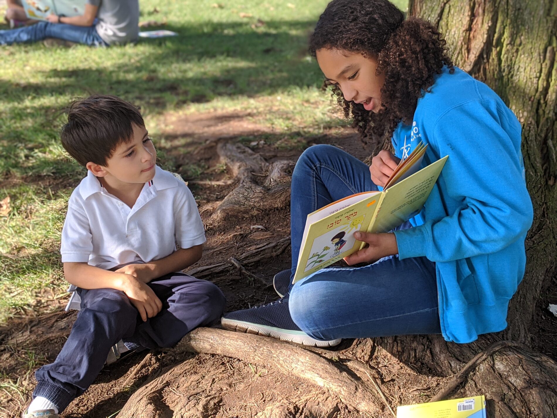 An older student at Kavod Charter School reads a picture book to a younger student. They are sitting outside together.