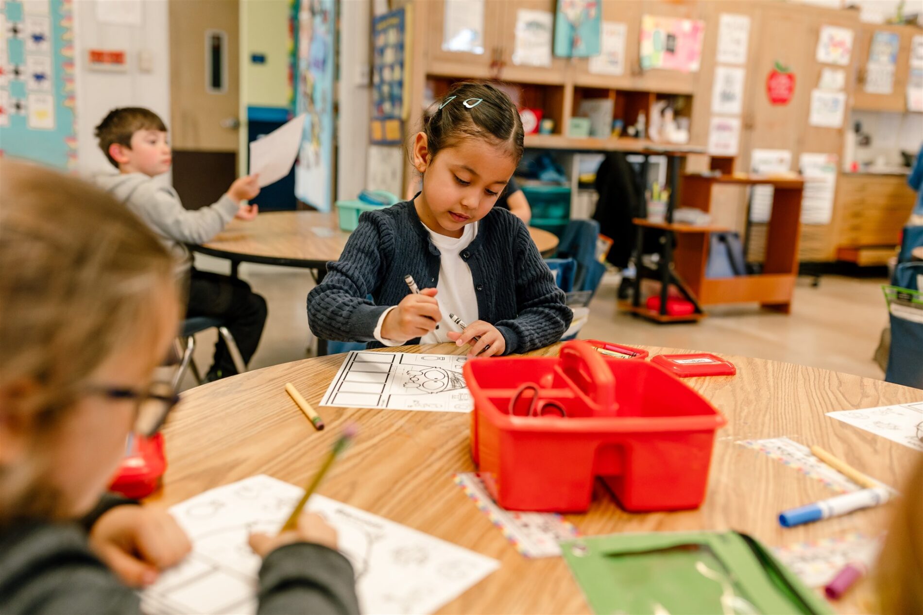 A Kavod student is focused and working independently at a collaborative table in a bright classroom. She is surrounded by classmates also working independently.