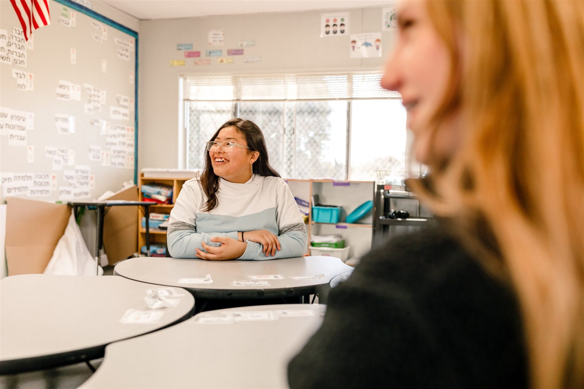 Kavod Middle School student sitting at a desk in a brightly lit classroom, while smiling and staring eagerly at the front of the room.