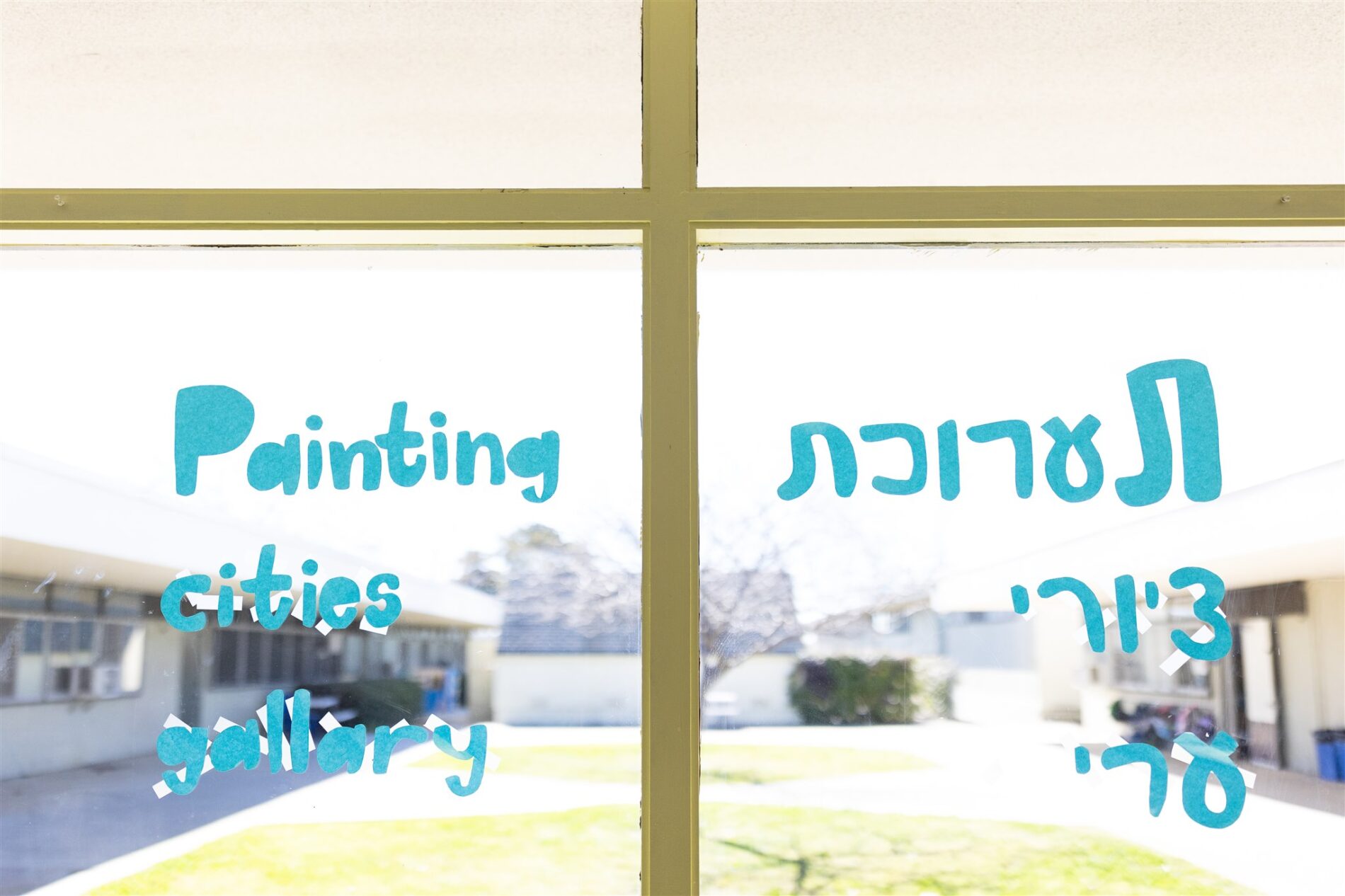 A glass window that overlooks the Kavod campus with the words “painting” “cities” and “gallery” in English and in Hebrew.