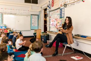 A Kavod Teacher sits in a chair at the front of the classroom, reading to students who are sitting on the carpet.
