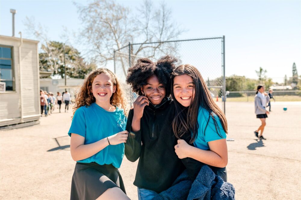 Three Kavod middle schoolers smile in the sunshine while gathered together outside of the school.