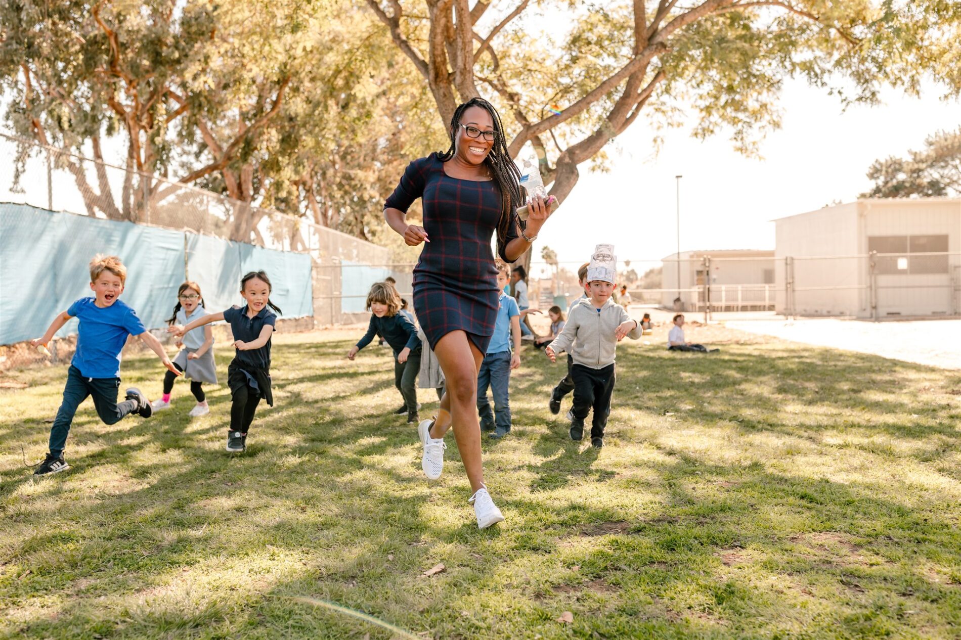 A Kavod Teacher smiles while jogging outside during recess while students chase closely behind her.