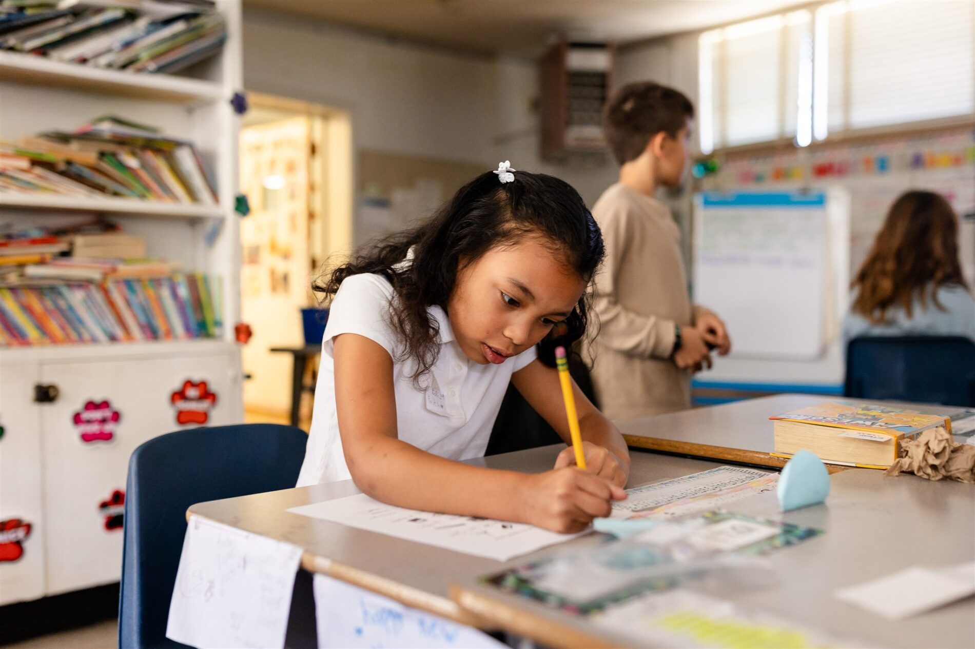 A Kavod student holds a pencil and attentively works on a worksheet at her desk.