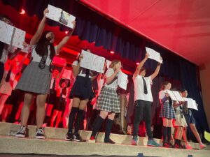 Two lines of Kavod Middle Schoolers stand on stage with bright red lights and a curtain behind them, holding up binders and notebooks during this year’s Matilda performance.
