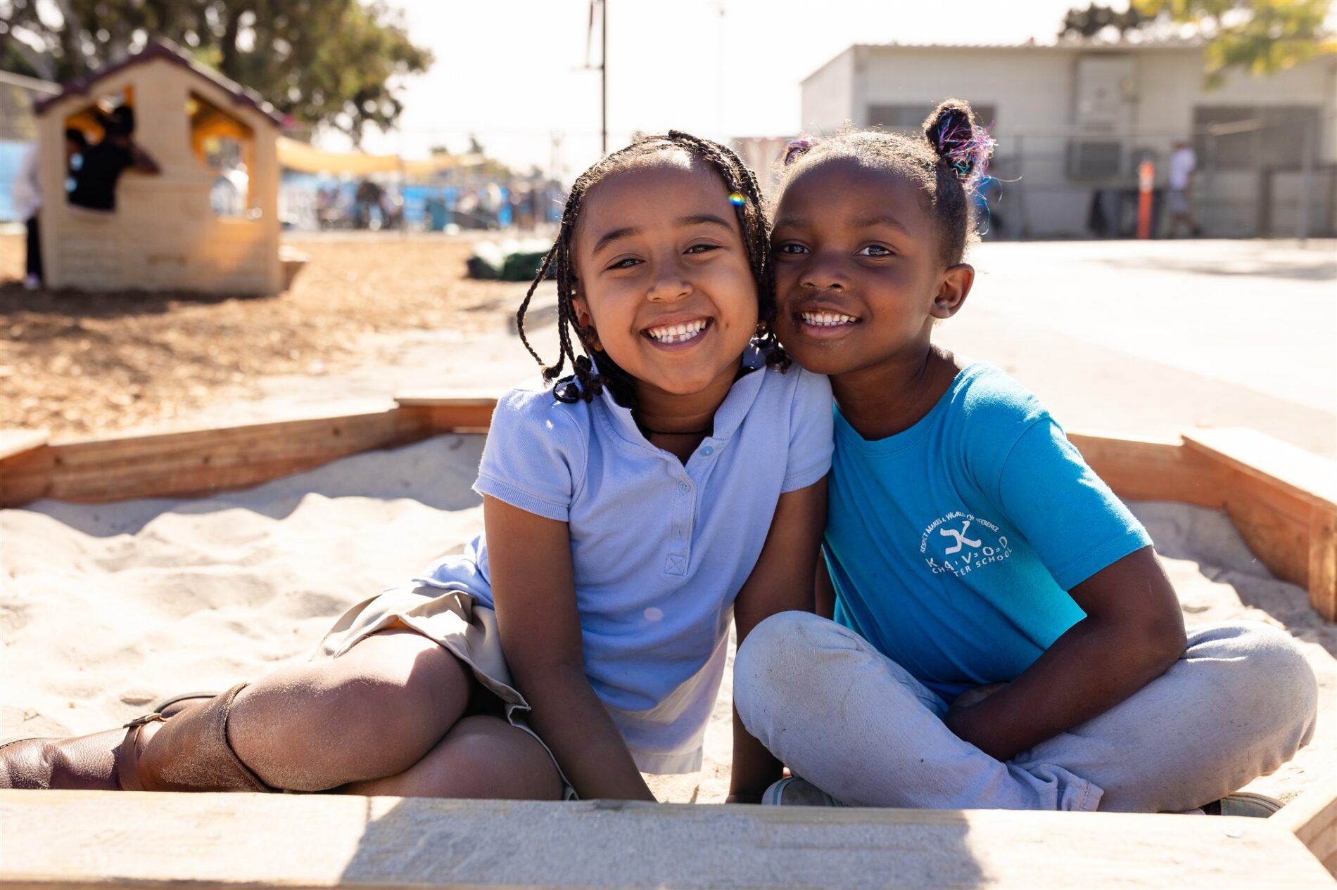 Two Kavod students sitting in a sandbox on a sunny day, smiling at the camera with their heads next to each other.