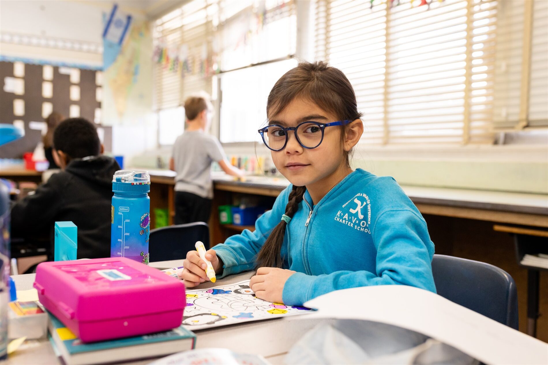 Kavod student in a Kavod zip-up sitting at a desk in the classroom while holding a marker stares at the camera.