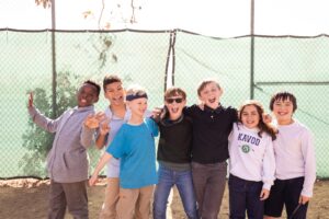 A group of 7 Kavod students stand outside smiling, mouths wide, huddled together in front of a fence.
