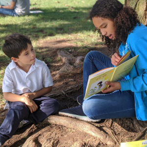 Two people sitting outside near a tree, one is holding open a book and reading a story in Hebrew to a child looking at the pages.  