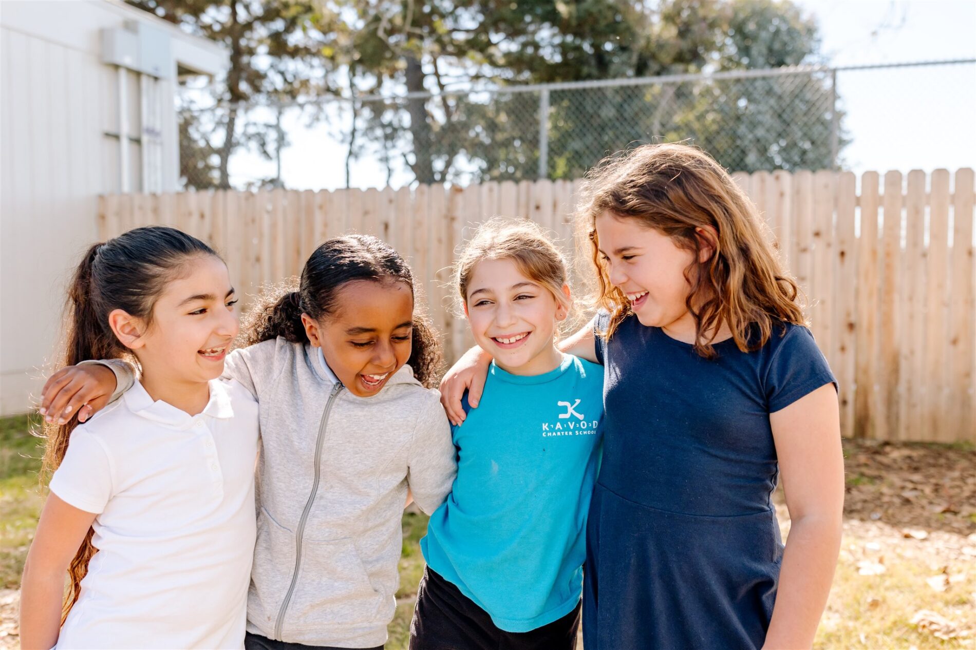 Four Kavod students stand together with arms linked smiling outside in front of a fence.