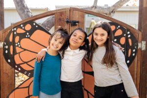 hree Kavod students stand together outside of Kavod’s Butterfly Garden smiling at the camera.
