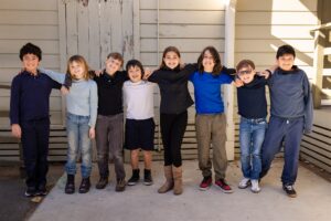 Kavod students lined up outside, standing shoulder-to-shoulder with their arms around each other, smiling at the camera.
