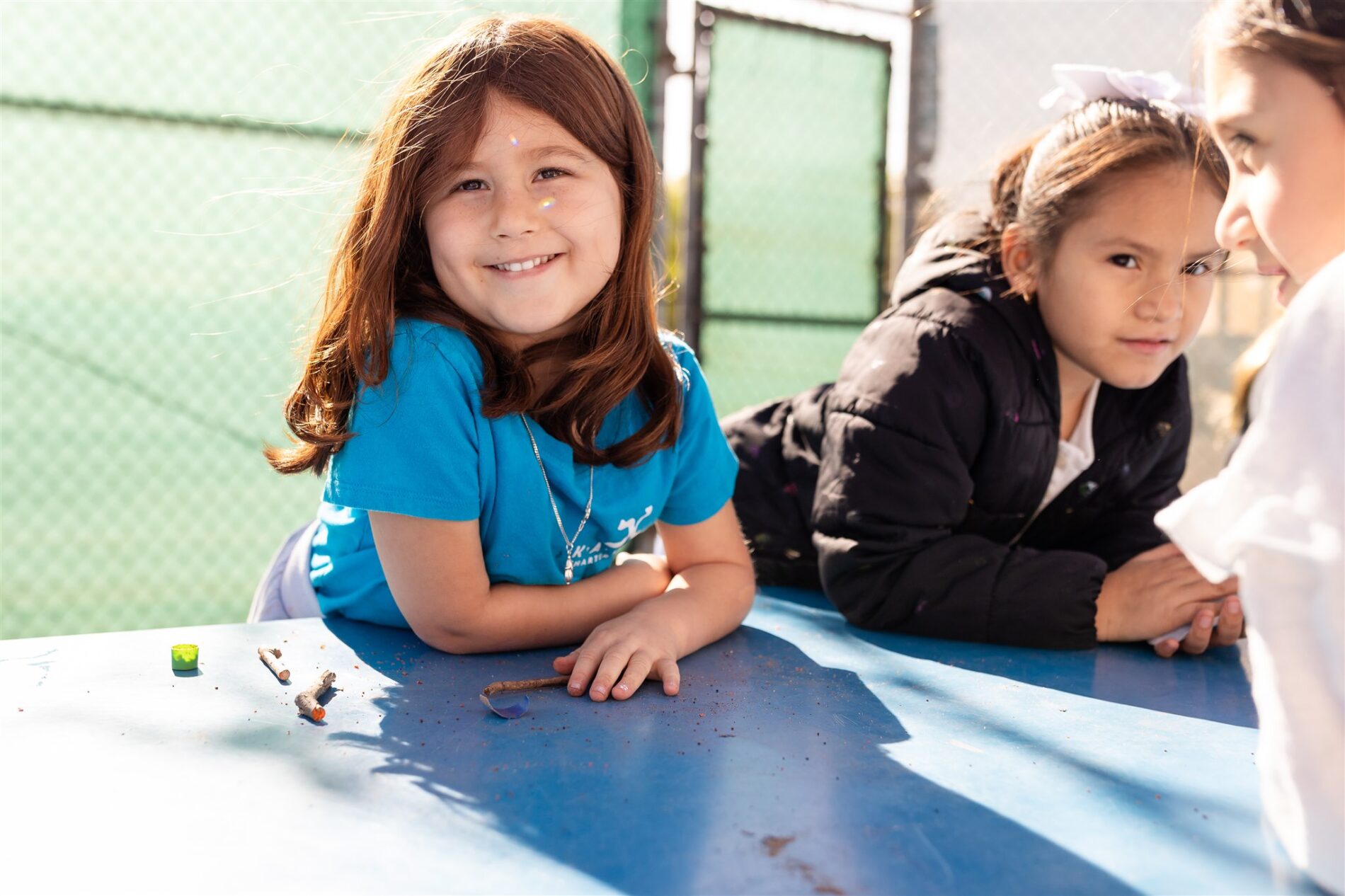 A Kavod student sits outside at a table, smiles at the camera as the sunshine makes rainbows on her face.