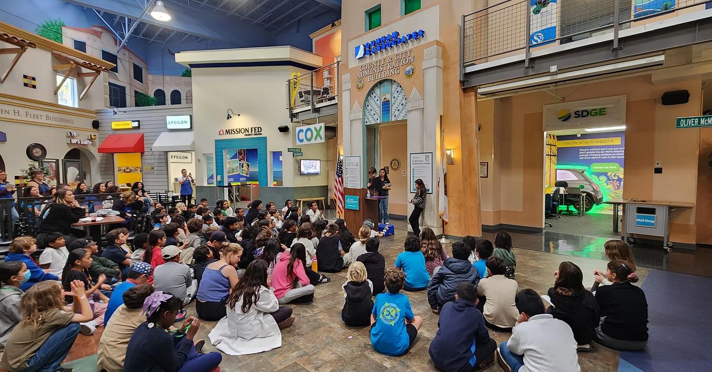 Kavod students at Biz Town, facing the County & City Administration Building, while students talk at a podium.