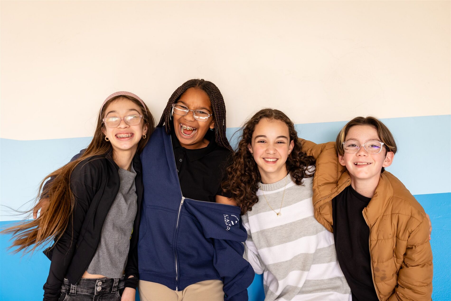 Four Kavod middle school students standing against a wall, smiling at the camera with their arms wrapped around each other. 