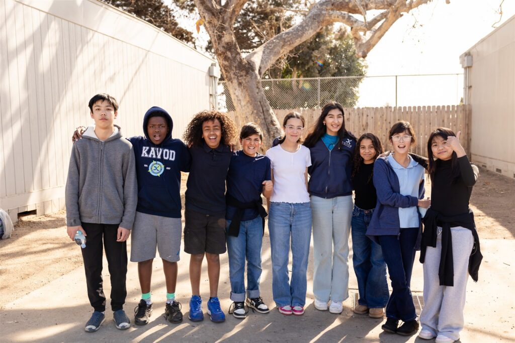 A group of Kavod middle school students stand outside with arms around each other, smiling at camera.
