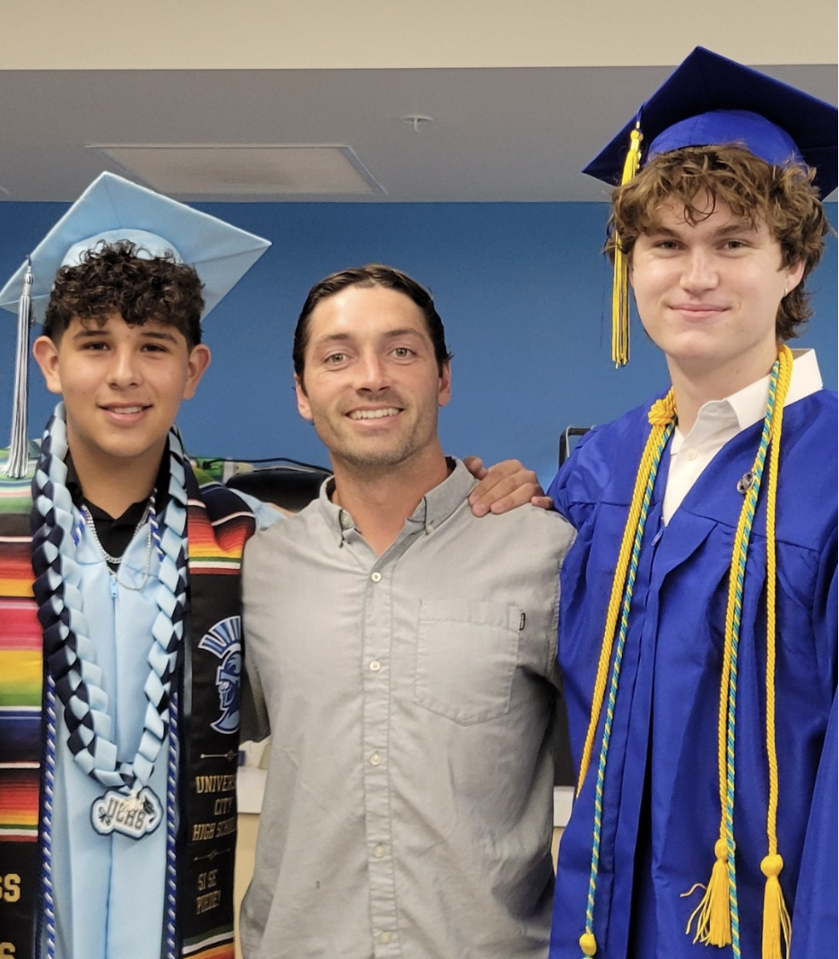 Mr. MacLeod stands in the middle between 2 students in their high school graduation cap and gowns.