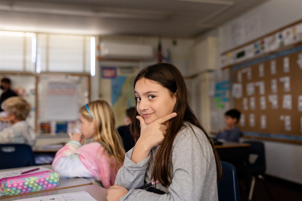 A Kavod student playfully smiles at the camera with her hand under her chin while in a classroom.